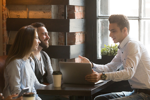 Insurance broker or salesman making offer to young millennial couple using laptop in cafe, realtor consulting customers about mortgage sitting at coffeehouse table pointing on computer screen